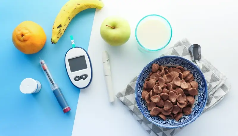 A table featuring a glucose meter, an insulin pen, a bowl of chocolate cereal, fruits (banana, orange, and apple), and a glass of milk, symbolizing a balanced approach to diabetes management through nutrition and monitoring.