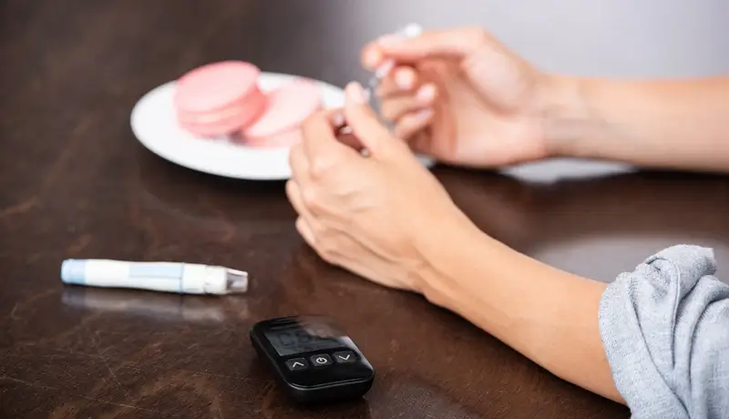 Hands preparing an insulin syringe on a table with a glucose meter, an insulin pen, and a plate of pink macarons, symbolizing the balance between diabetes management and dietary indulgence.