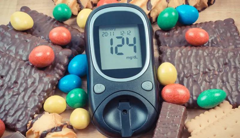 A glucose meter displaying 124 mg/dL surrounded by chocolates, biscuits, and colorful candies, highlighting the challenges of managing blood sugar levels while consuming sugary foods.