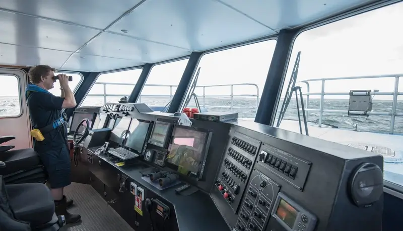 Ship officer on the bridge using binoculars to scan the horizon, with navigation systems and ocean view in the background.