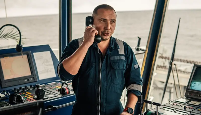Ship captain speaking on a radio device in the vessel's control room with navigation equipment in the background.