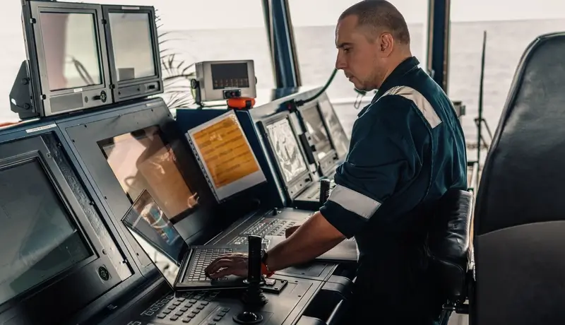 Ship officer operating navigation systems on a vessel's bridge, surrounded by multiple monitors and controls.