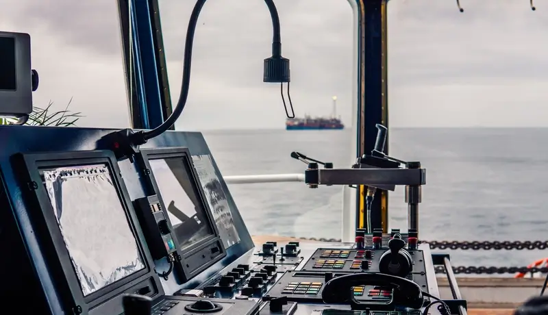 Control panel on a ship's bridge overlooking the sea with another vessel in the distance.