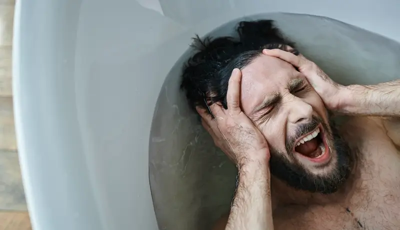 A distressed man in a bathtub holding his head with both hands, screaming in apparent pain or frustration.