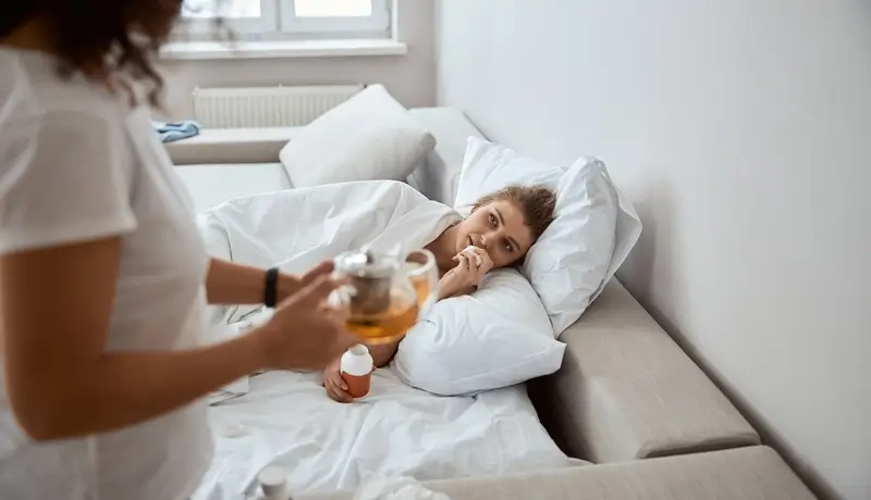 A sick woman lying on the couch while someone brings her tea