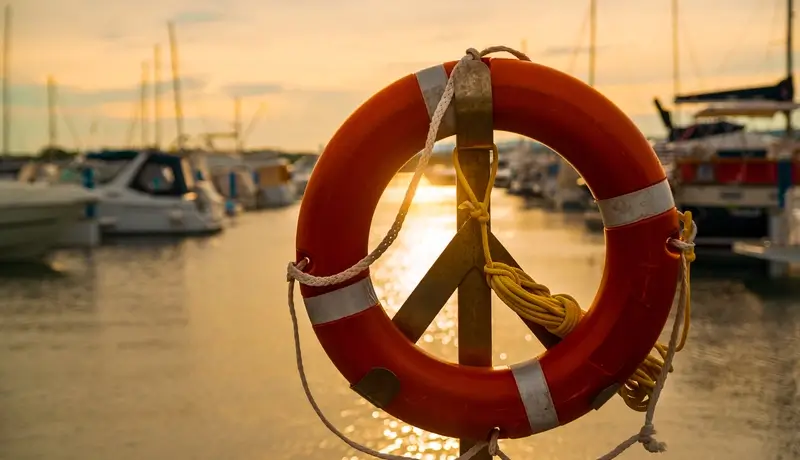 A lifebuoy hanging on a pole in a marina at sunset, with boats and a peaceful water scene in the background.