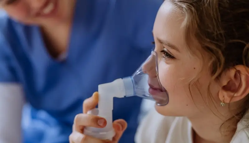 Little girl with inhaler in hospital room, nurse checking her.