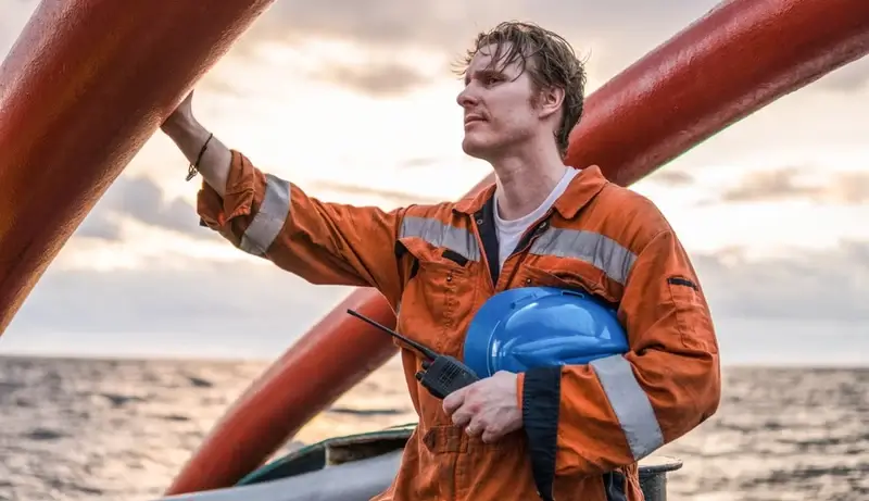 A seafarer in an orange safety suit holding a blue helmet on a ship's deck, gazing thoughtfully into the distance at sea.