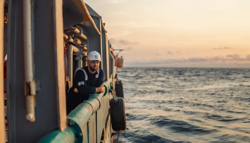 A sailor wearing a helmet gazes at the sea from the deck of a ship as the sun sets on the horizon.