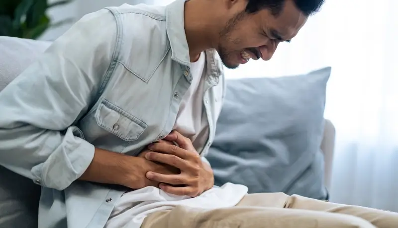 A man clutching his abdomen in severe pain, sitting on a couch, possibly experiencing symptoms of abdominal distress.