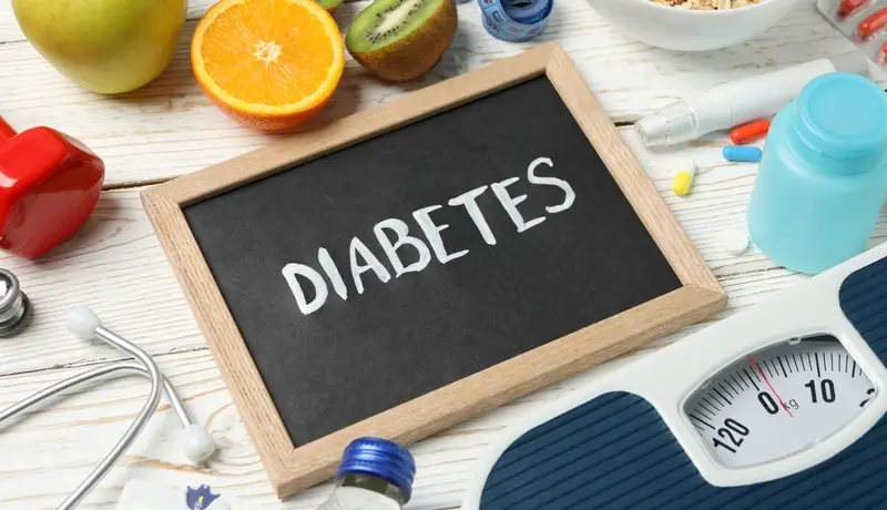 Μια μαύρη πινακίδα με τη λέξη "Diabetes" περιβάλλεται από φρούτα, ιατρικά εργαλεία και μια ζυγαριά, συμβολίζοντας τη διαχείριση του διαβήτη.
